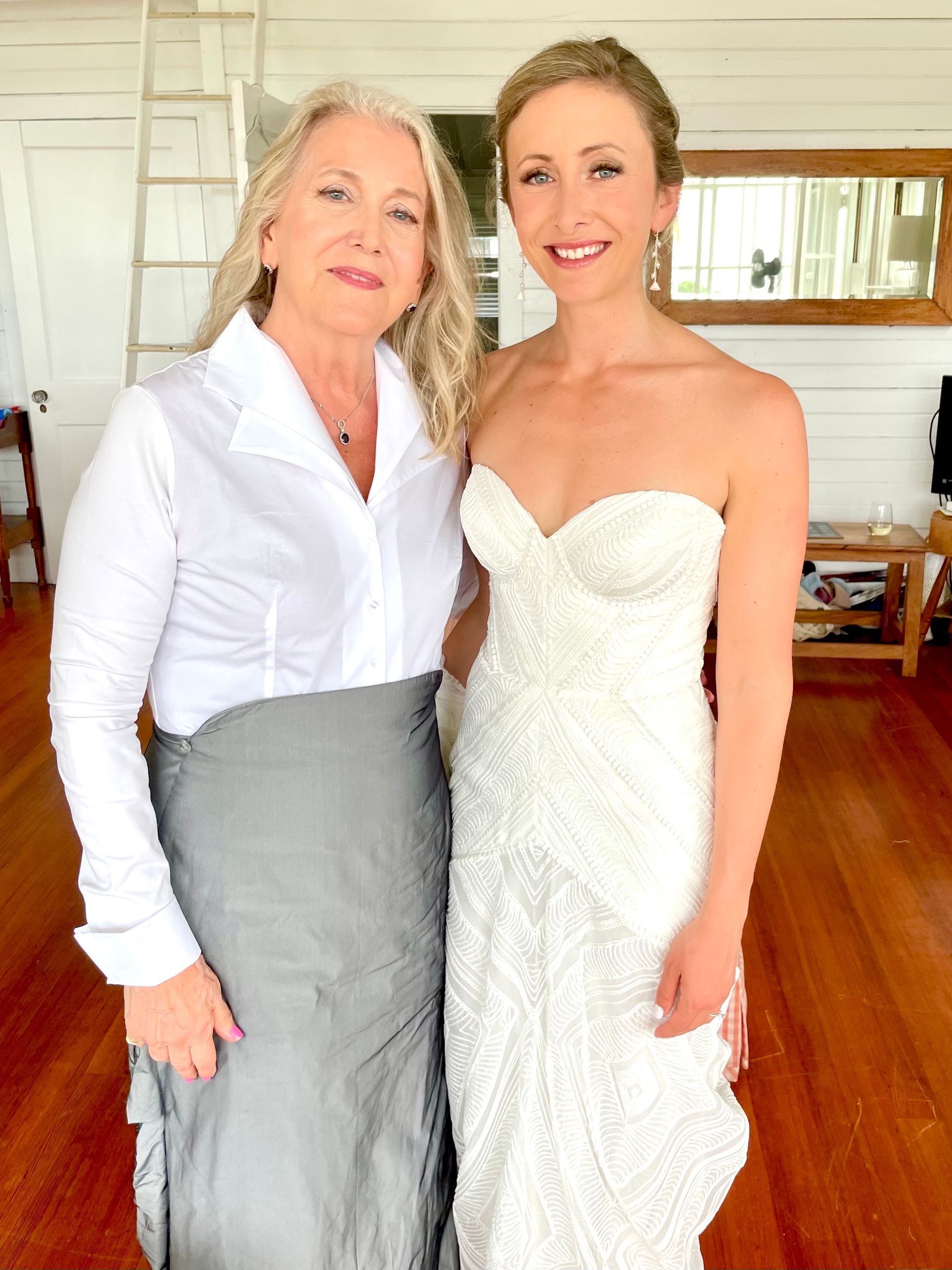 Two women standing together indoors, one in a white blouse and gray skirt, one in a white strapless dress.