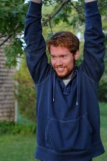 Man in a navy hoodie smiling with arms raised outdoors in a yard