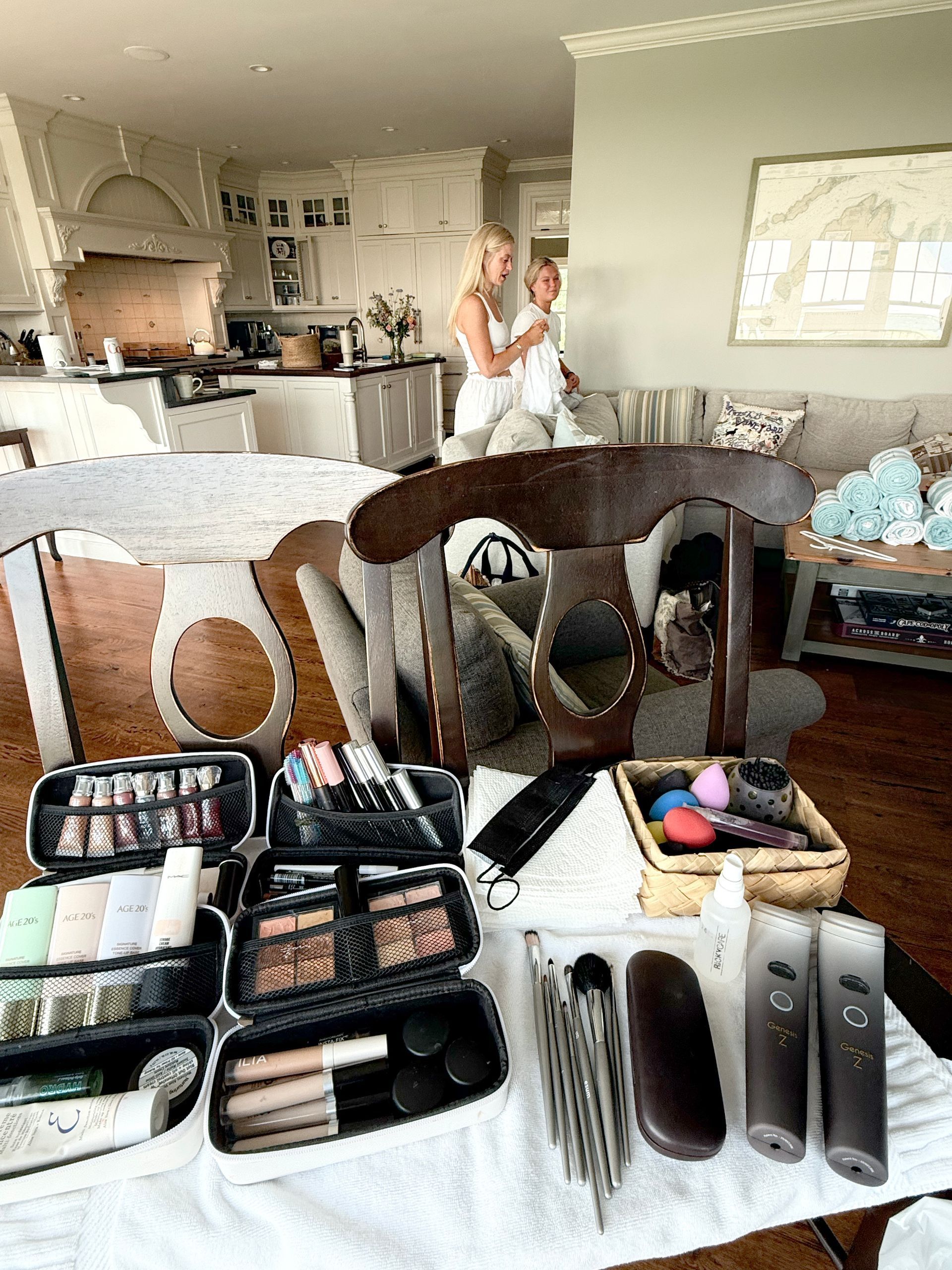 Makeup and cosmetics spread on a table in a bright living room, with a woman standing in the background.