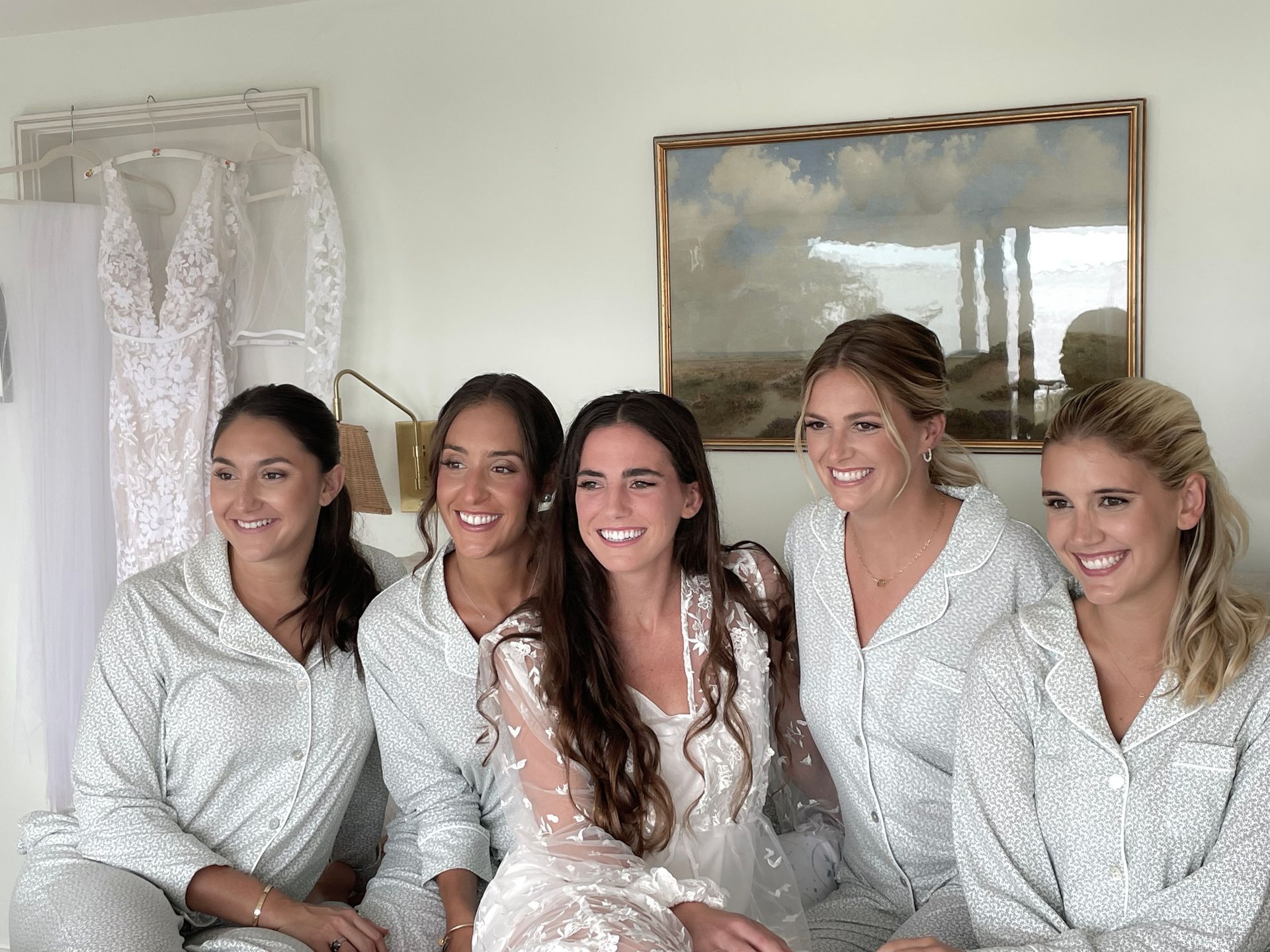 Five women in white robes smiling together indoors on a bed, with a framed mirror behind them.