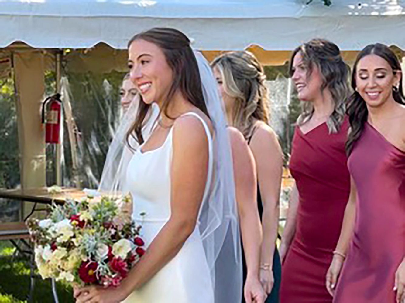 Bride in white veil holding a bouquet, smiling beside bridesmaids in burgundy dresses at an outdoor wedding