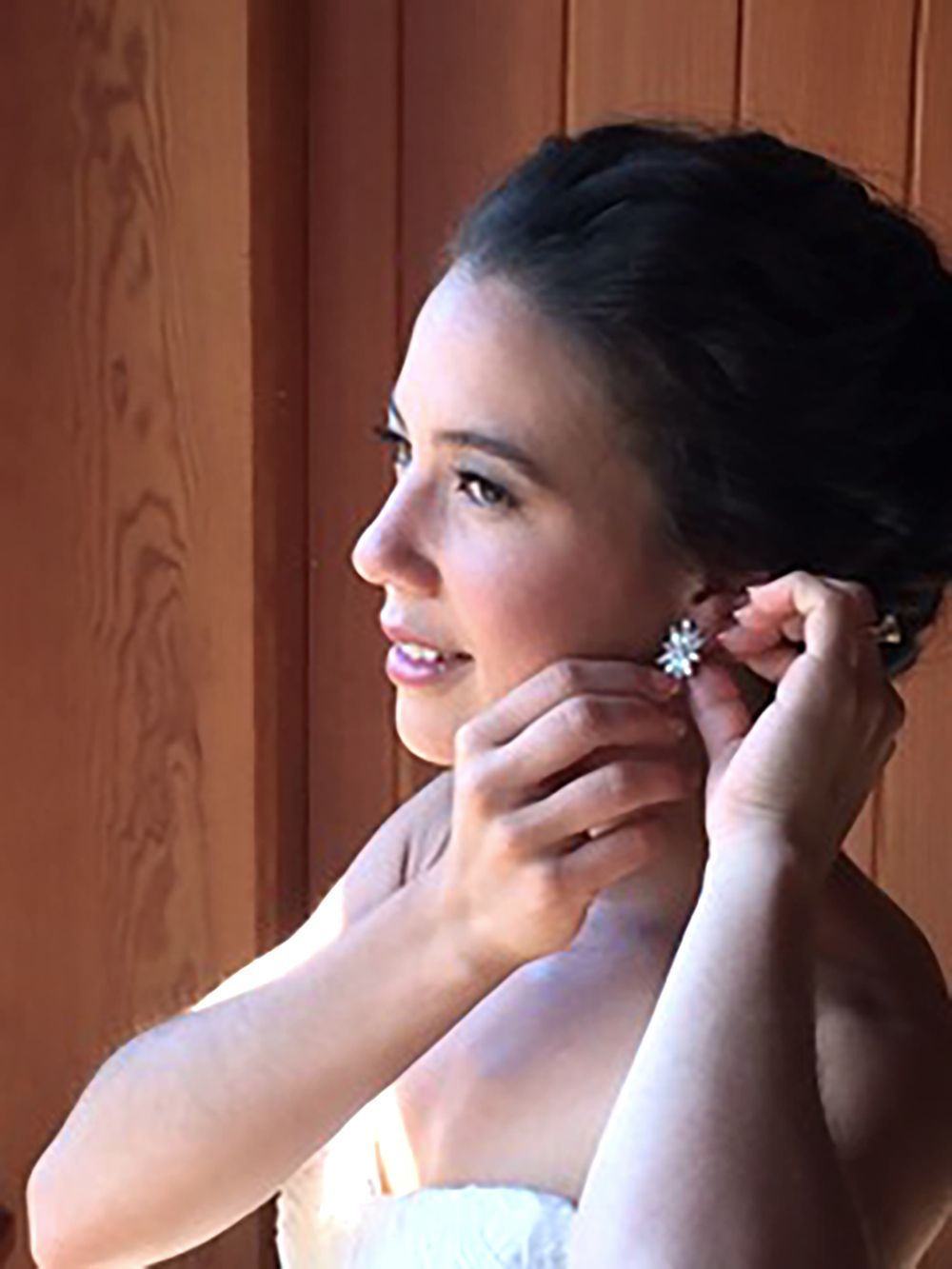 Woman putting on an earring by a wooden wall, side profile with a slight smile.