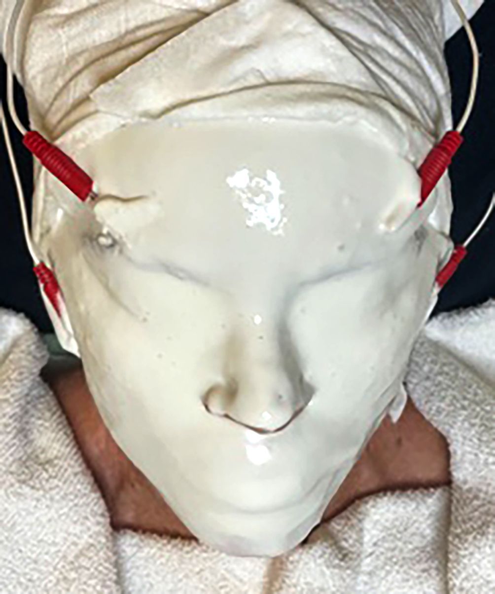Close-up of a white theatrical mask with red side straps and a cloth wrap around the head