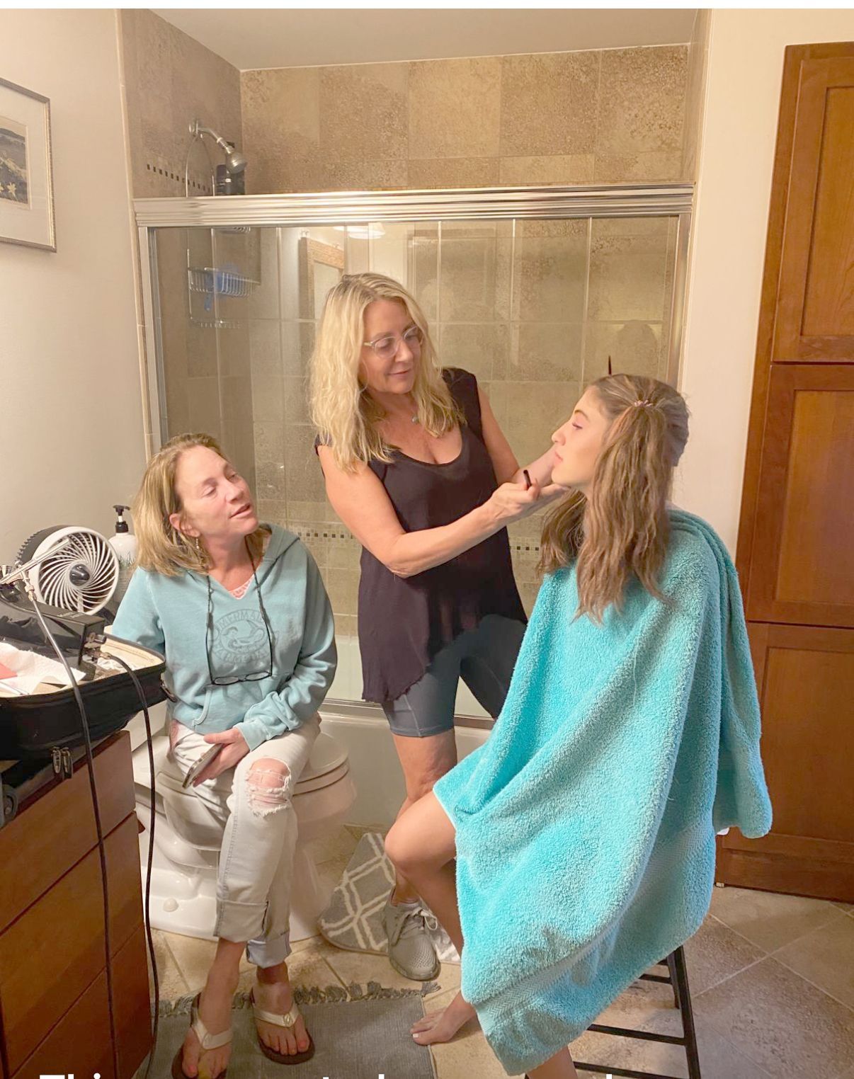 Three women in a bathroom, one styling another's hair while a third sits nearby.