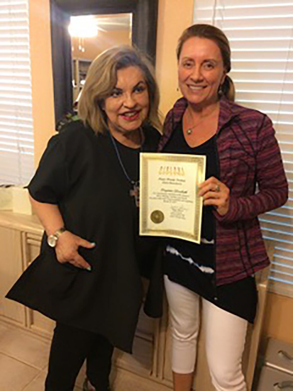 Two women smiling and holding a certificate indoors возле a doorway