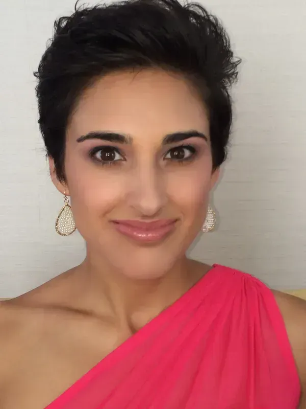 A person with short, dark hair wearing a bright pink one-shoulder top and ornate gold earrings, smiling at the camera.