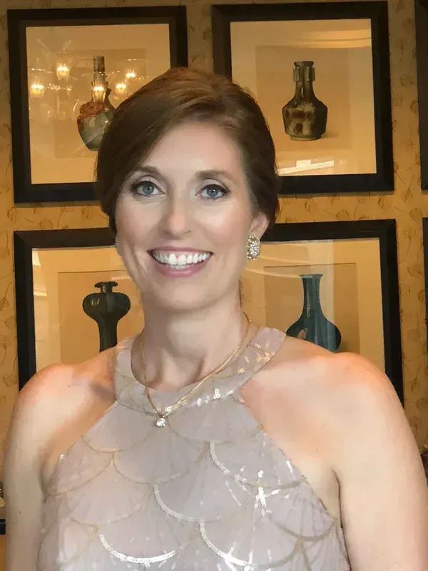 A smiling person wearing a light pink, scalloped-pattern top stands in front of a wall decorated with framed art prints.