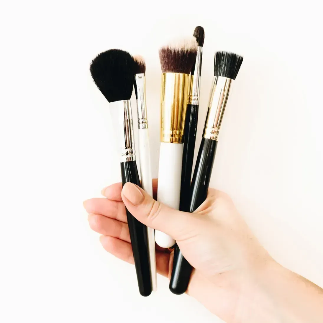 A hand holding a cluster of five various cosmetic makeup brushes against a white background.