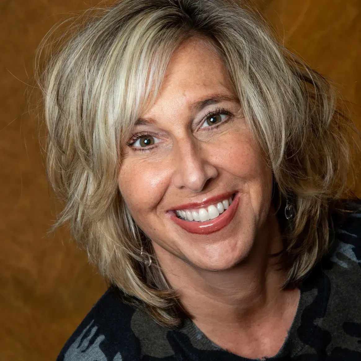 A woman with shoulder-length blonde hair smiles warmly at the camera against a textured, dark gold background.