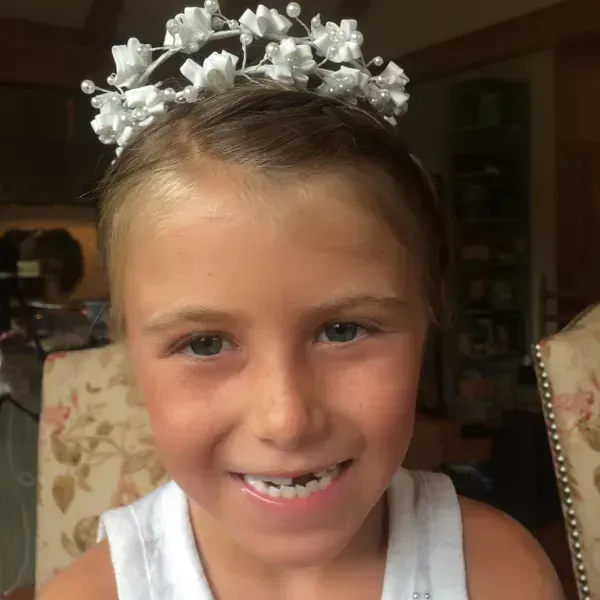A smiling person wears a white floral crown with small, pearl-like accents, set against a blurry indoor background.