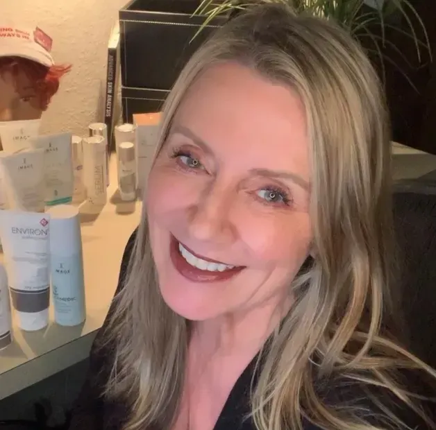 A smiling blonde woman looks toward the camera with several skincare product bottles displayed on a desk in the background.