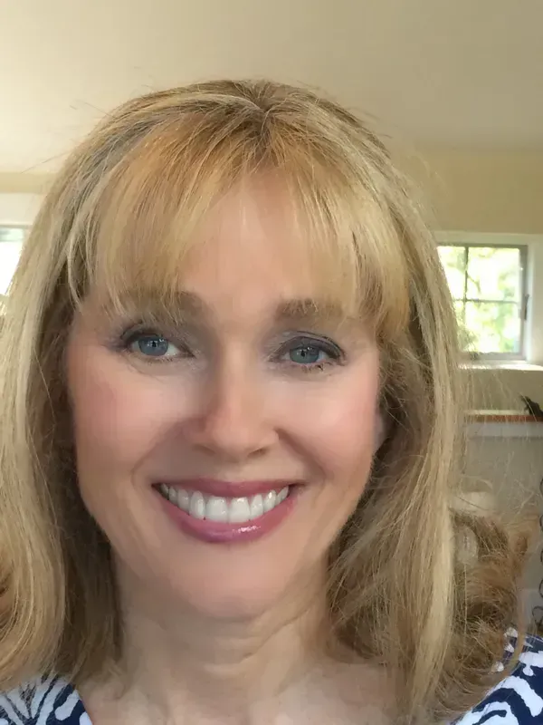 A smiling person with blonde hair and bangs, wearing a blue and white patterned shirt, indoors near a window.