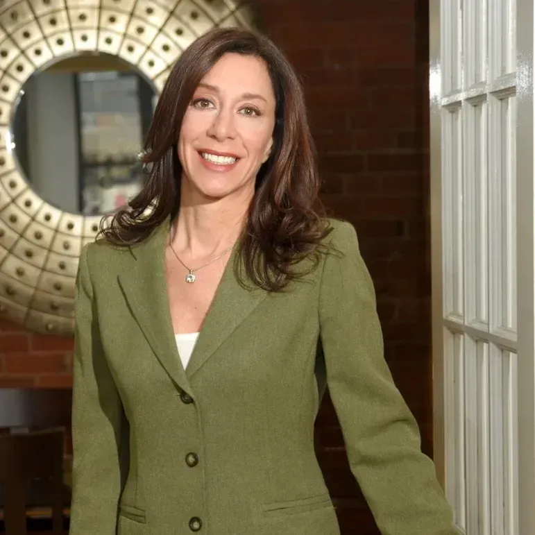 A smiling professional wearing a green blazer, standing in an office setting with a decorative wall mirror.