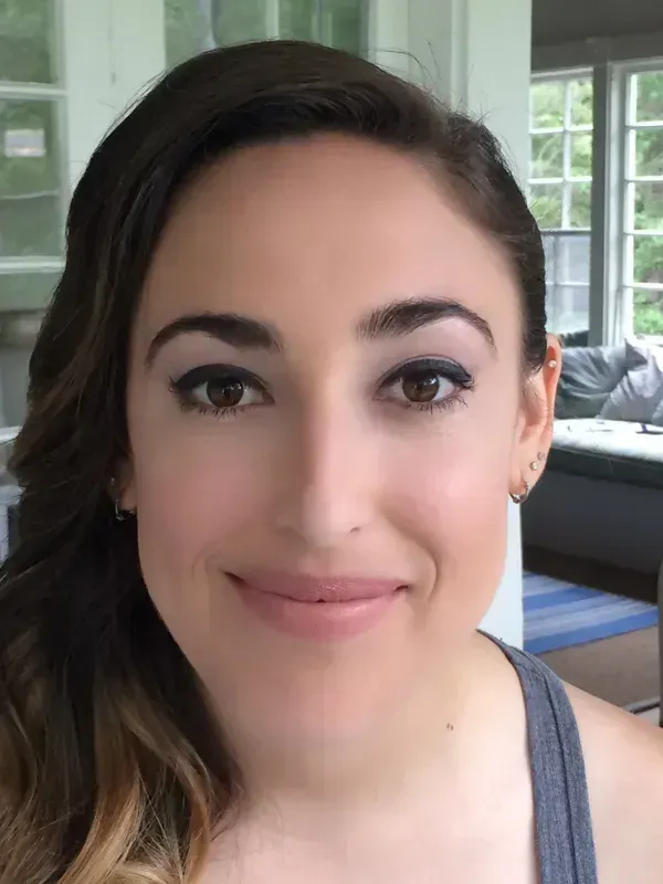A person with shoulder-length hair smiling warmly at the camera in a room with windows in the background.