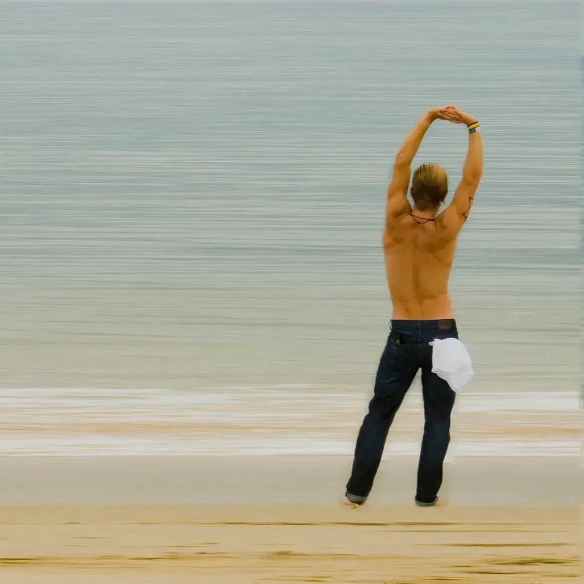 A shirtless person stands on a sandy beach, facing the ocean with their arms stretched high above their head.