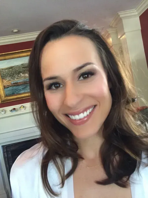 A smiling person with shoulder-length brown hair, wearing a white robe in a brightly lit room with a framed painting.