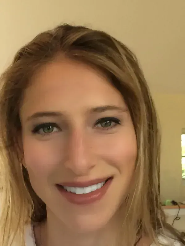 A close-up portrait of a smiling person with blonde hair, looking directly at the camera in a brightly lit room.
