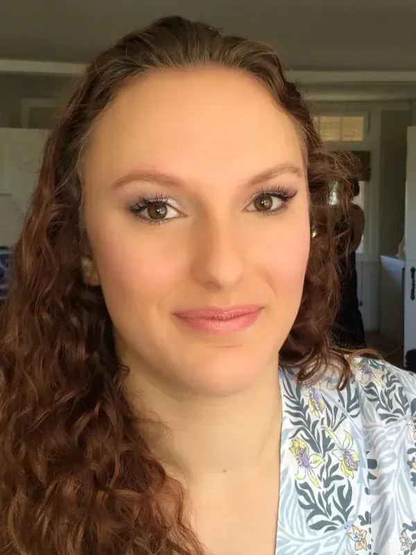 A person with long, curly auburn hair and a neutral expression looking directly at the camera.