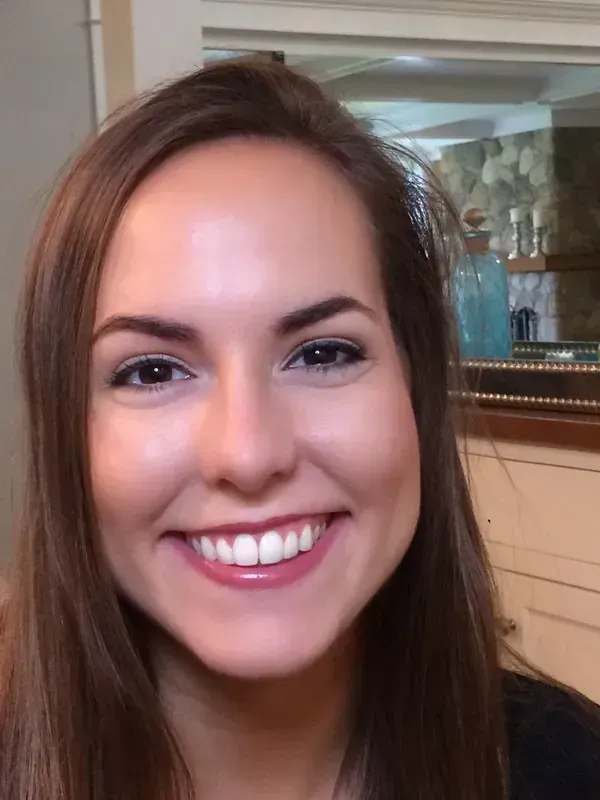 A person with shoulder-length brown hair smiling at the camera, set against an indoor home background.