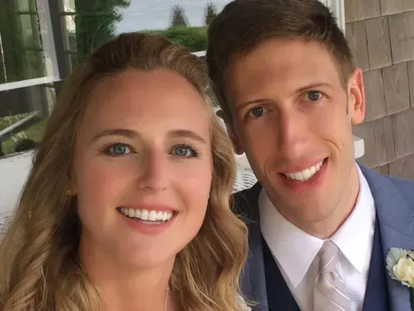 A smiling couple posing for a selfie outdoors in front of a shingled building.