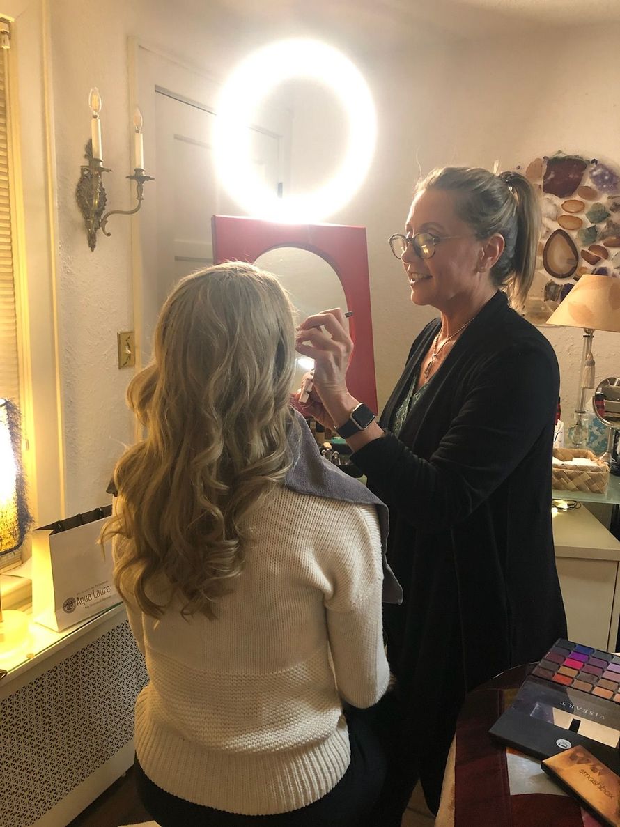 Woman doing makeup on another woman in a warmly lit room with a ring light and mirror