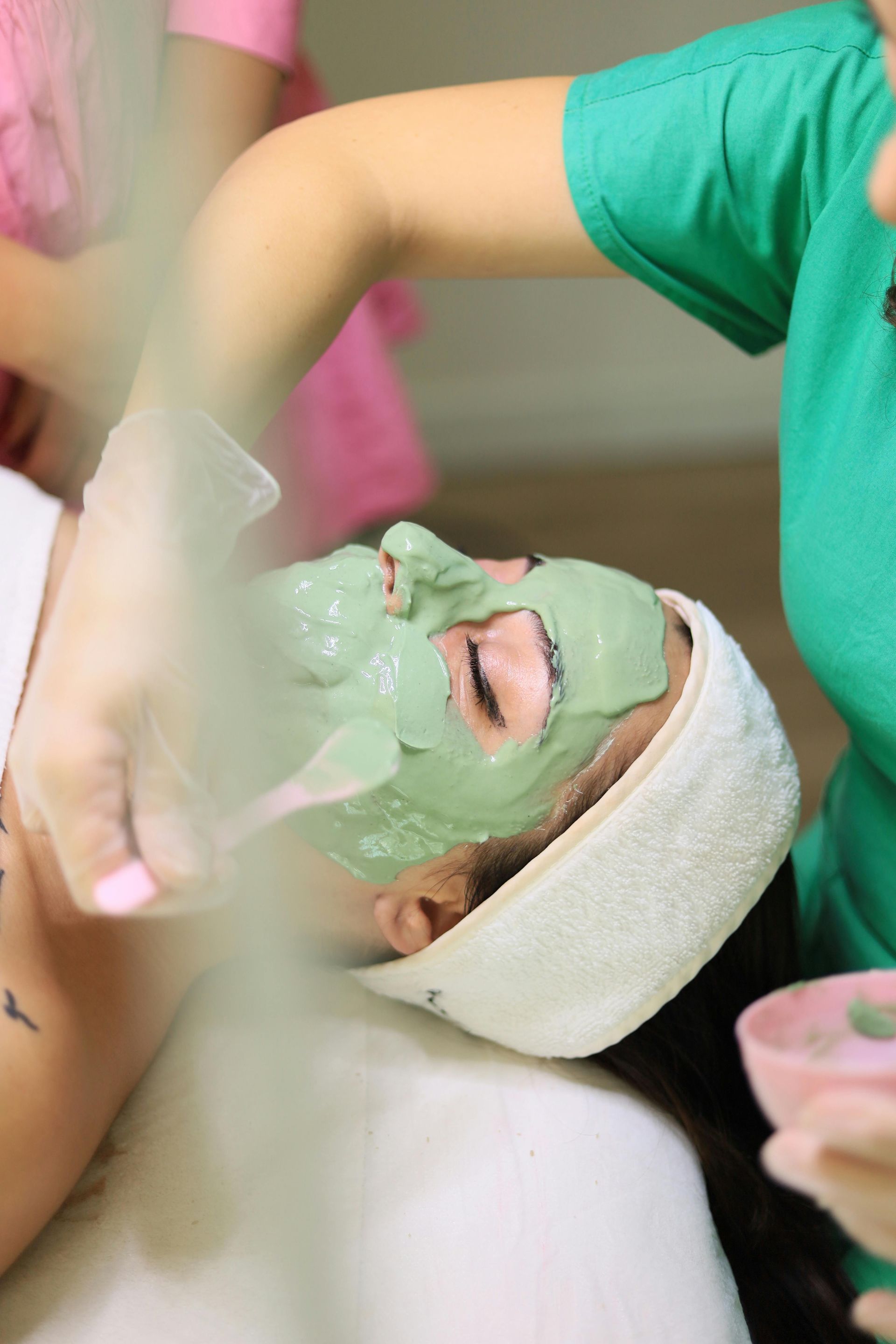 A professional in a green top applies a green facial mask to a client wearing a white headband in a spa setting.