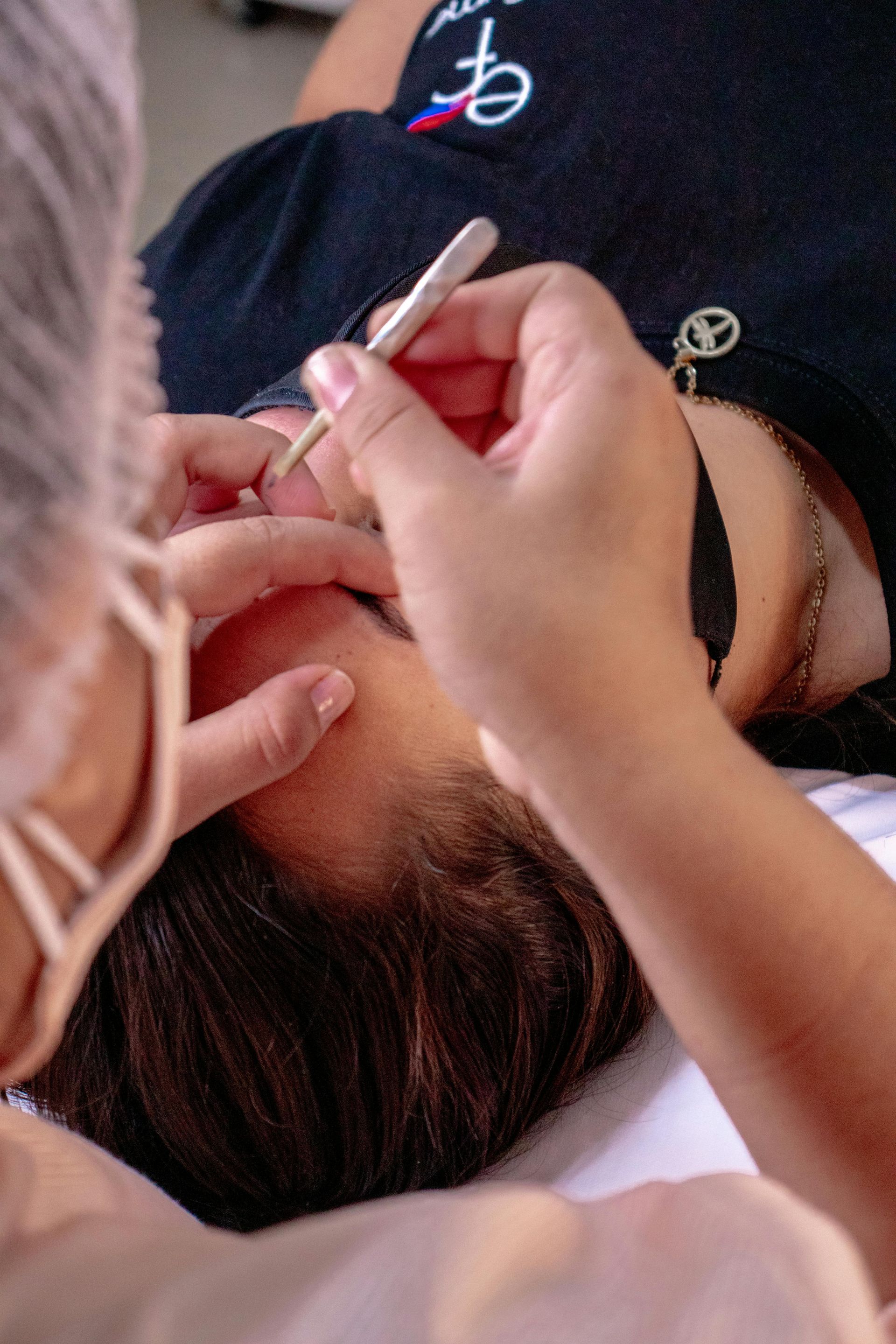 A person wearing a face mask and head covering uses tweezers to apply eyelash extensions to a client lying on a table.