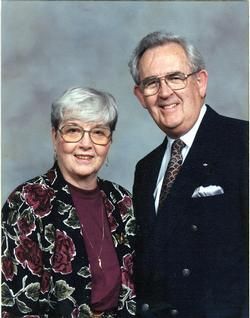 Smiling couple posing in formal attire against a studio backdrop, with the woman in a floral jacket and the man in a suit.