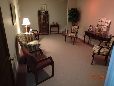 Empty waiting room with chairs, table lamp, side table, and brochure stand on beige carpet.