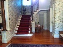 Interior hallway with a red-carpeted staircase, wooden floor, floral wallpaper, and a small table near the landing