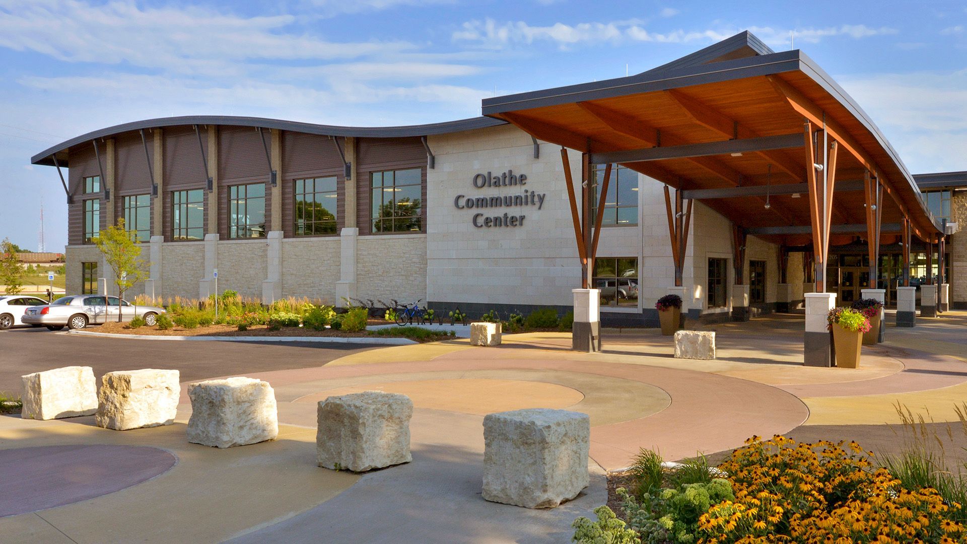 Olathe Community Center exterior, tan stone building, brown wood canopy and accents, curved entrance, blue sky.