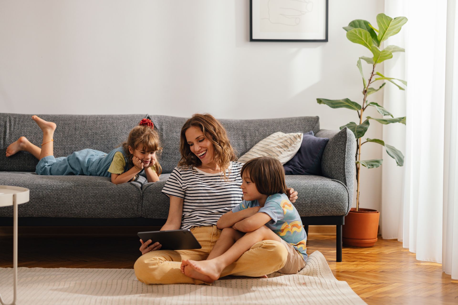 Woman and two children on a living room rug, looking at tablet. Sofa and plant in background.