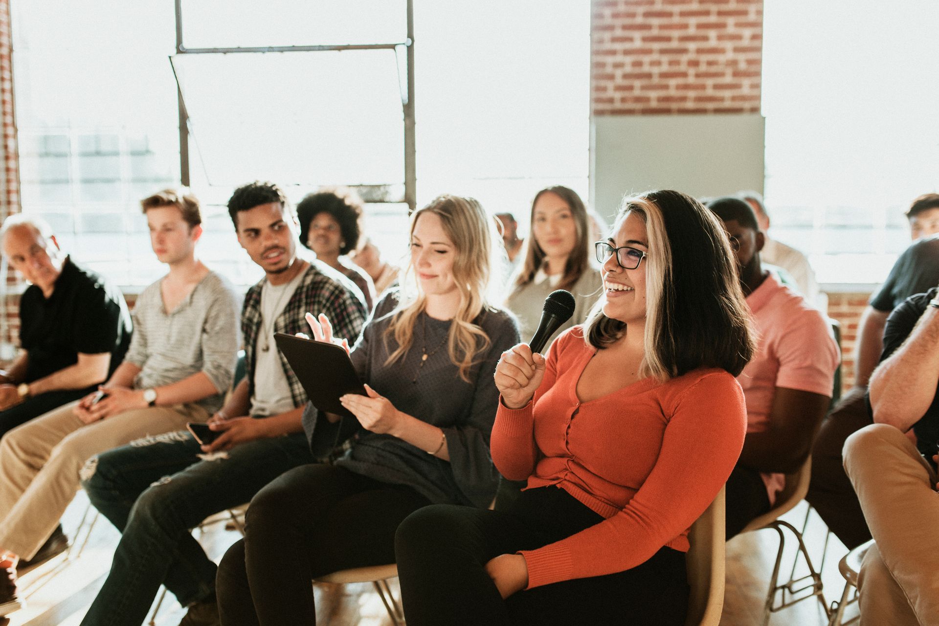 Woman with microphone smiles while speaking to seated audience.