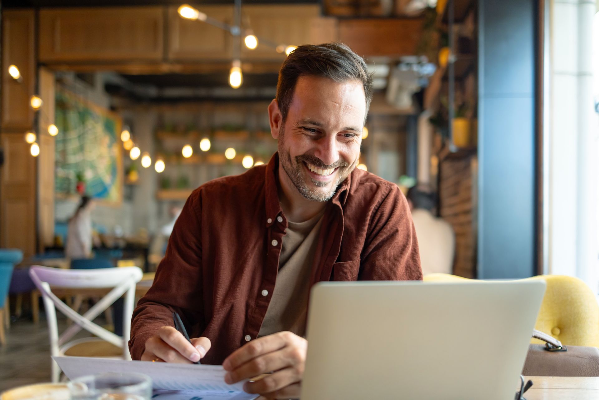 Man smiles while working on laptop and papers in a cafe.
