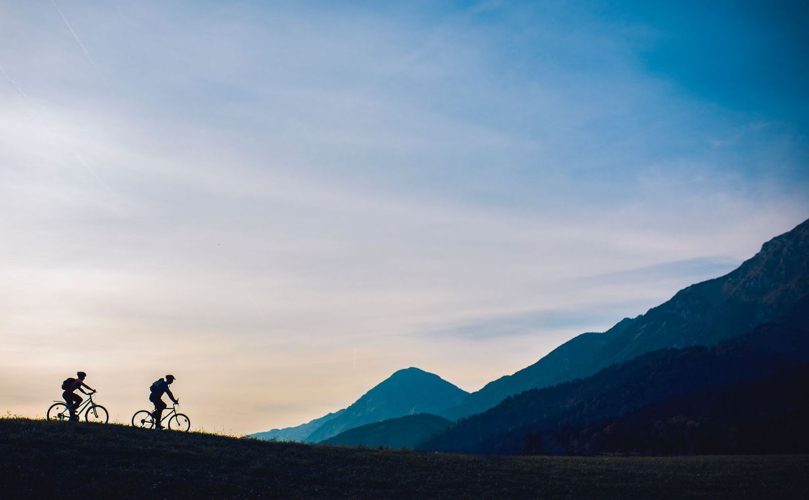 Two cyclists ride across a hilltop in front of silhouetted mountains against a cloudy sky.