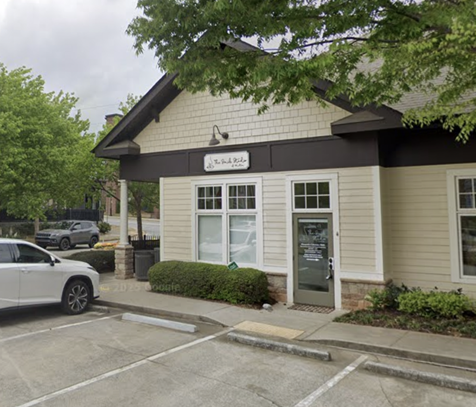Small cream storefront with dark trim and a glass door in a parking lot, partly shaded by trees