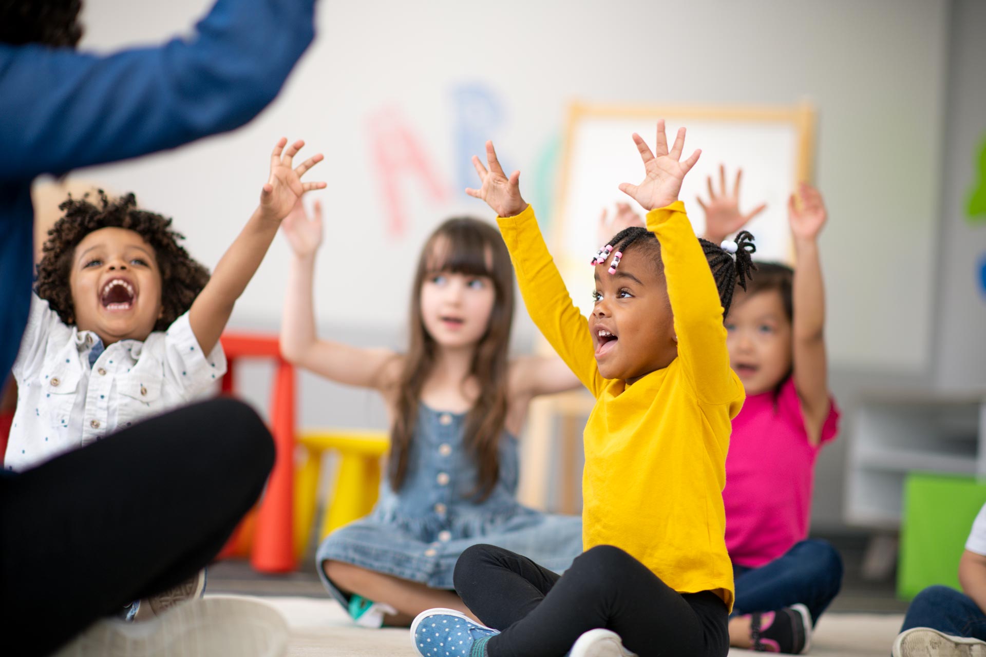 Children in a classroom with arms raised, led by a teacher.