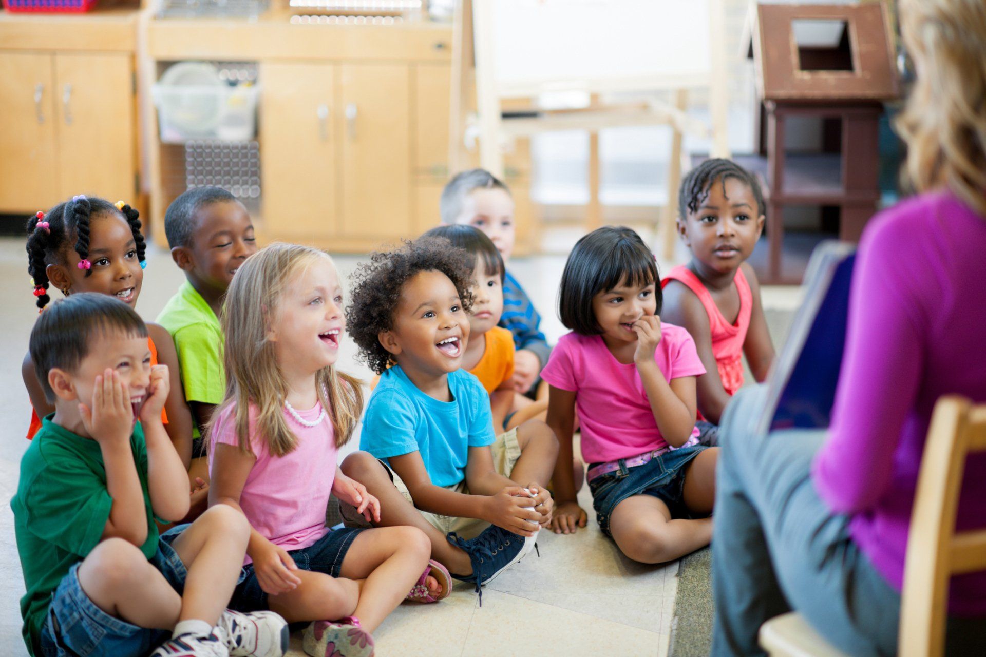 Children sitting on the floor, listening attentively to a teacher reading a book in a classroom.