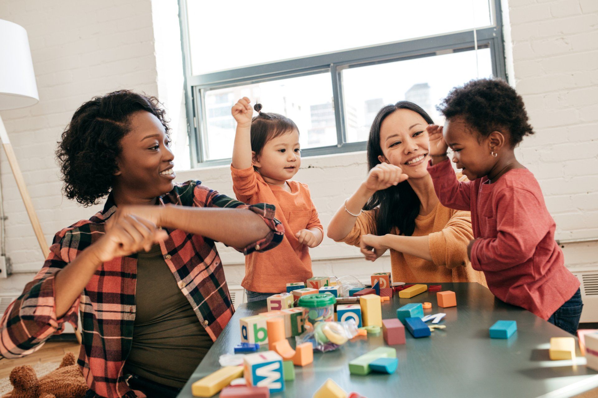 Two adults and two children playing with blocks at a table indoors, bright room.