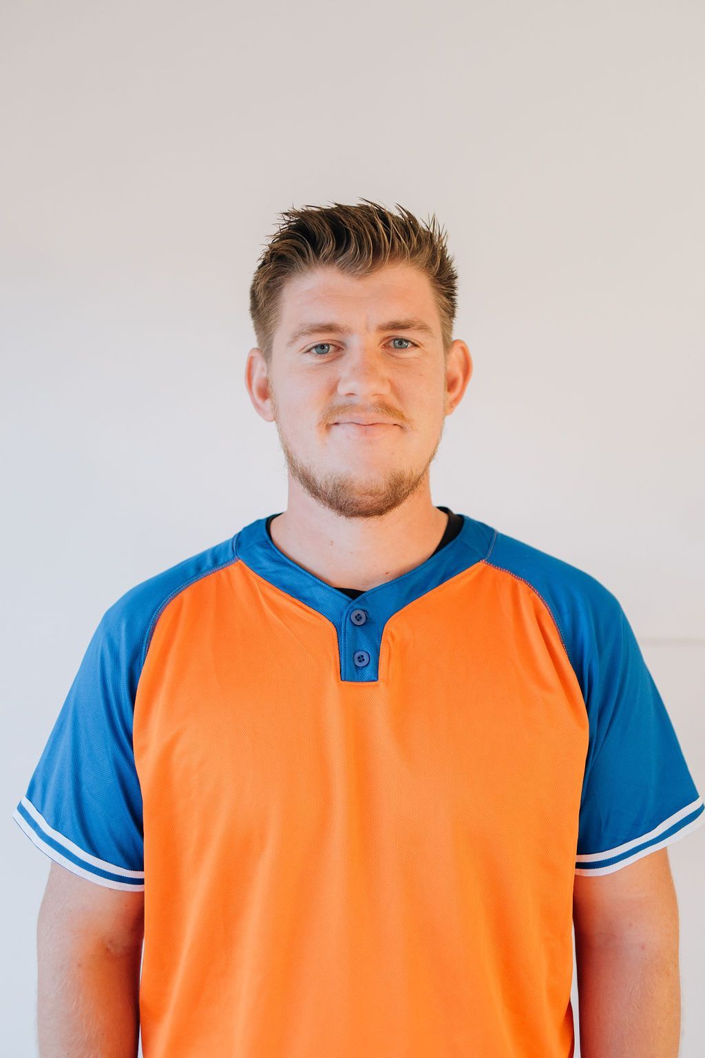 A man in an orange and blue shirt is standing in front of a white wall.