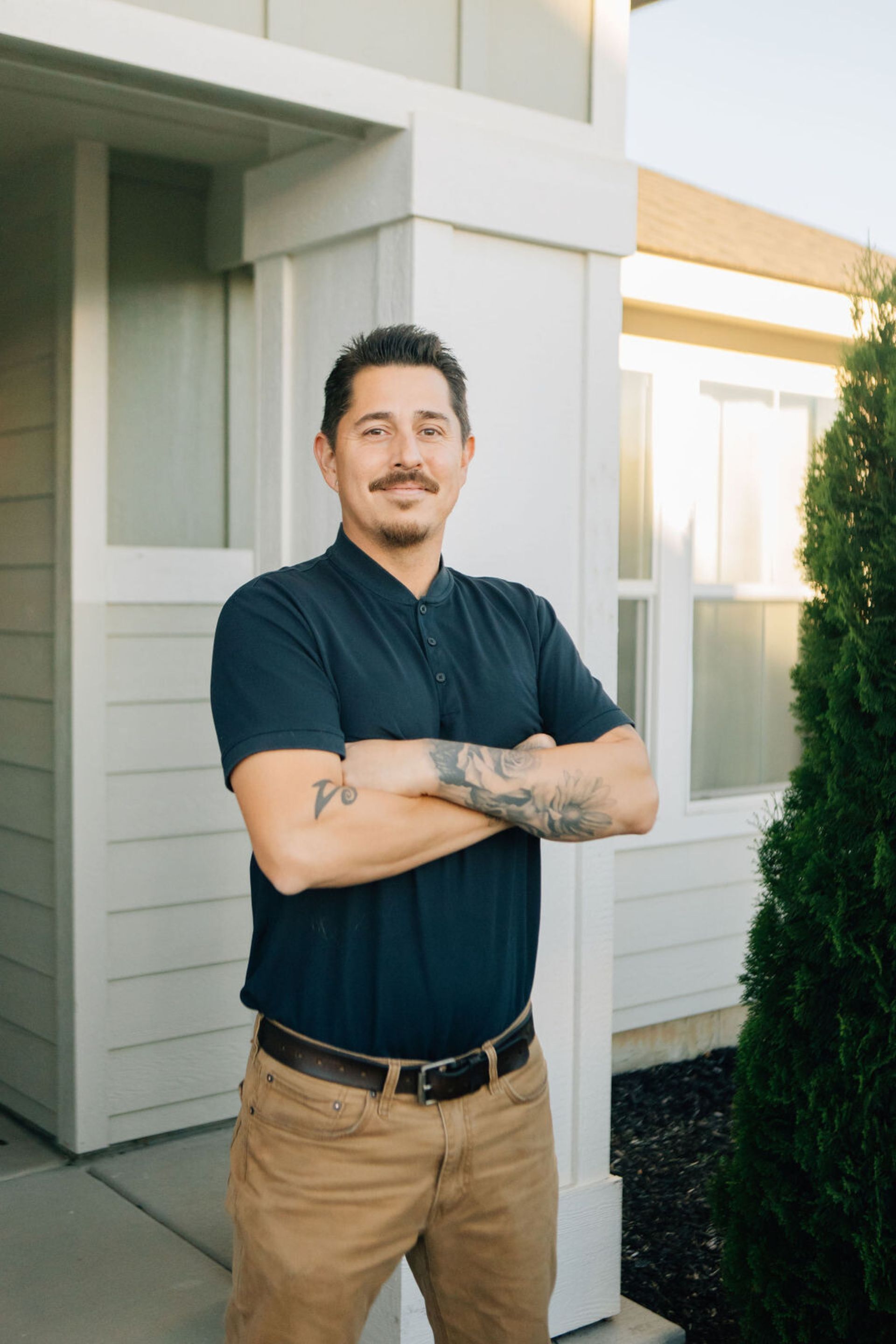 A man with a mustache is standing in front of a house with his arms crossed.