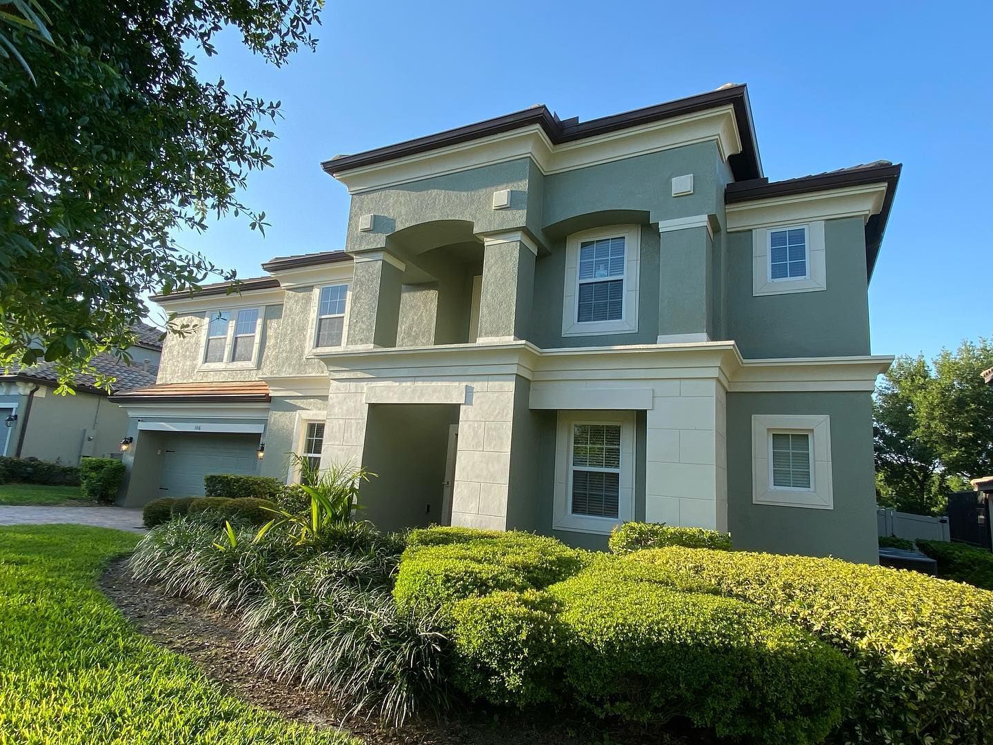 Two-story stucco house with green trim, white windows, and manicured hedges in front.