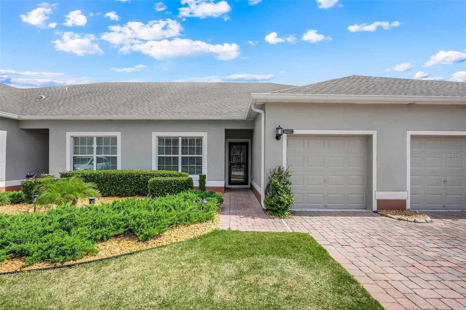 Single-story townhouse with white garage doors, front lawn, and brick walkway under a blue sky
