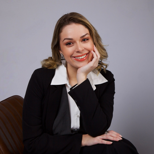 Mulher sorridente com blazer preto e camisa branca, sentada diante de um fundo cinza