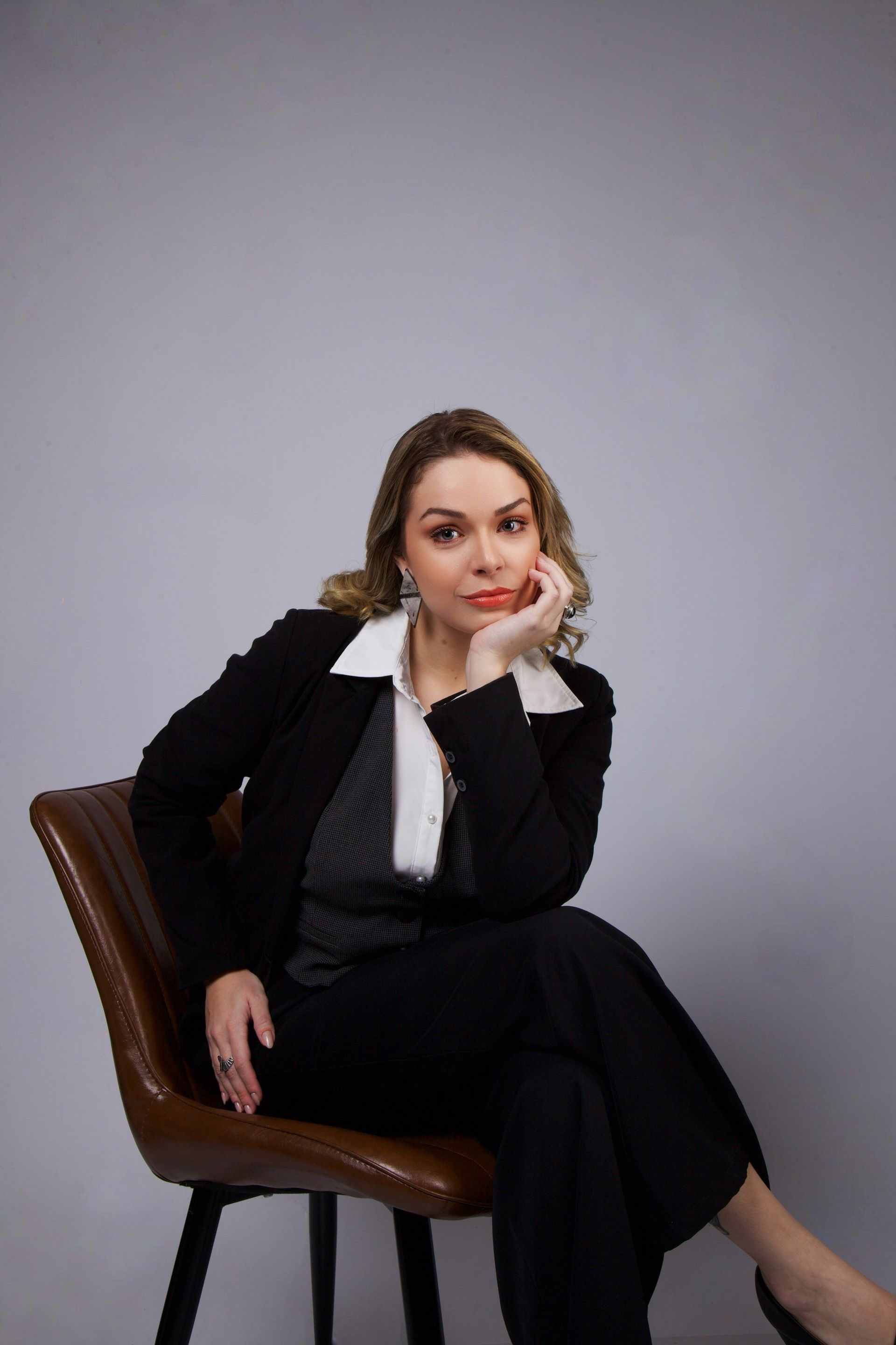 Mulher vestida de preto sentada numa cadeira marrom, com um fundo cinza liso, apoiando o queixo na mão