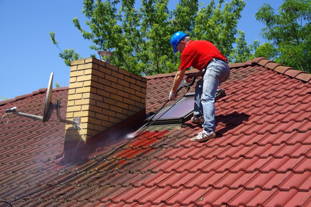 Person Pressure Washing a Red Tile Roof, Near a Brick Chimney and Satellite Dish — Ericson Constructions in Mareeba, QLD