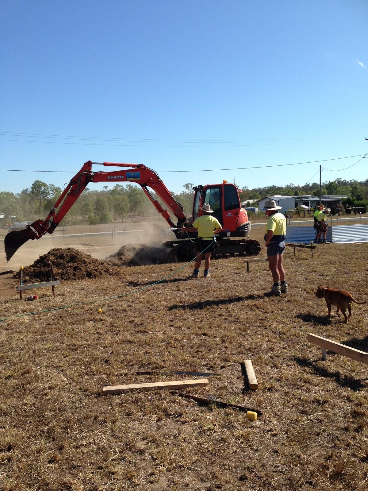 Excavator digging dirt at a construction site; two workers watching; dog nearby; sunny day.