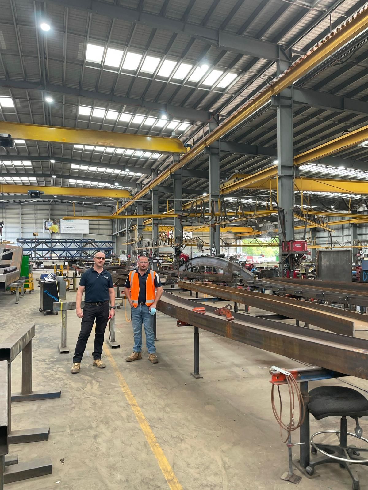 Two men in a factory, standing near metal beams. Overhead cranes, industrial setting.