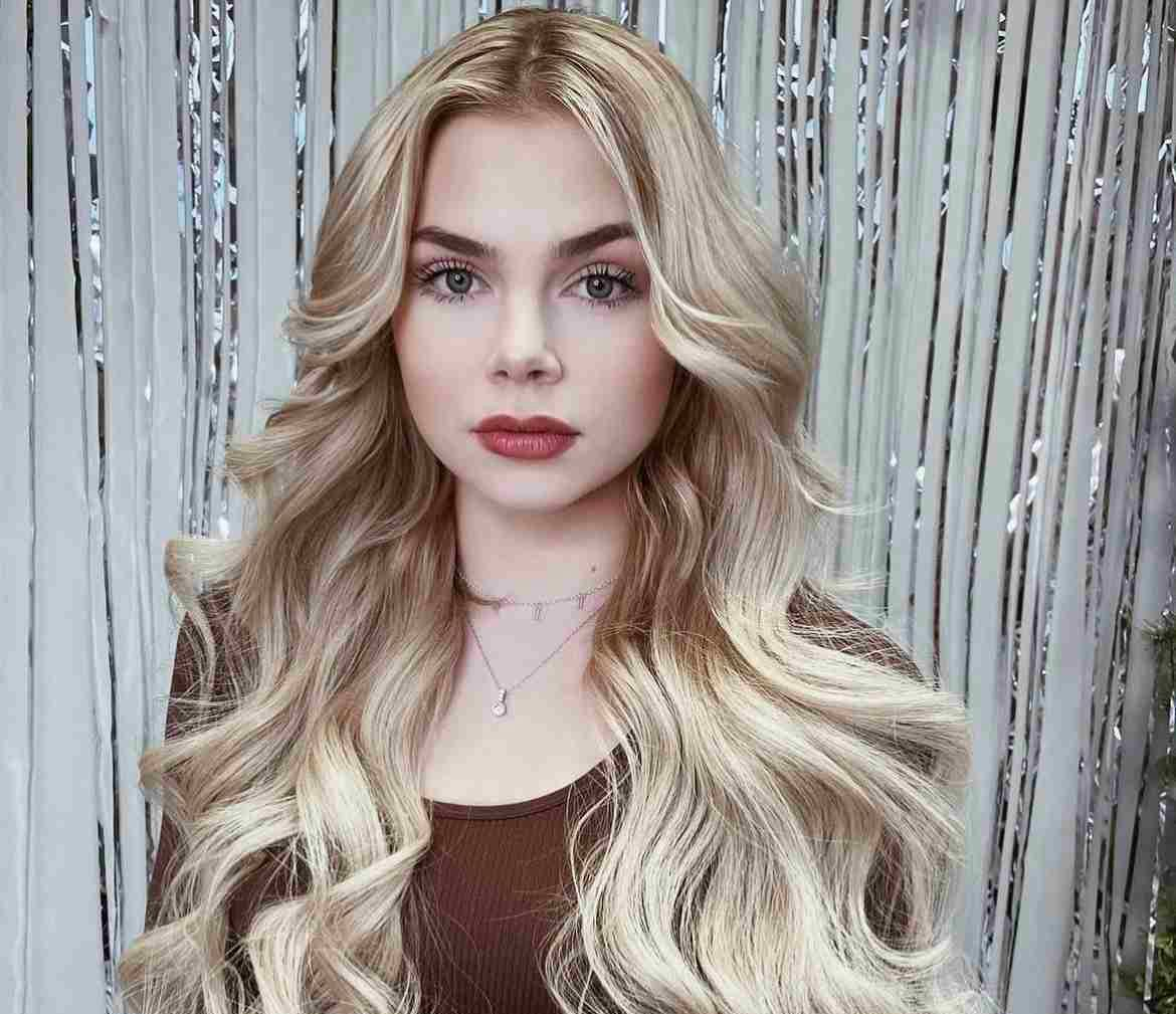 Woman with long, wavy blonde hair wearing a brown top and necklace, posing in front of a silver curtain.