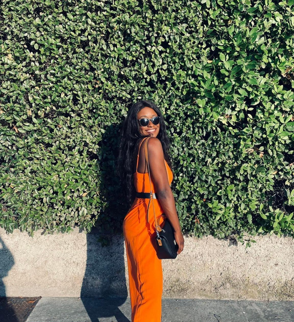 Woman in orange jumpsuit and sunglasses stands in front of a green hedgeld